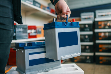 A person holding an automotive battery by its blue handle above another battery on a white surface. Shelves with car batteries and tools visible in the background