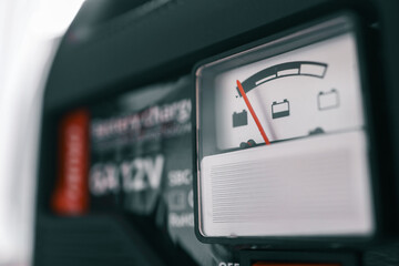 Close-up view of a battery charger gauge with focus on the voltage indicator. The red needle points to the current battery level, with blurred background details