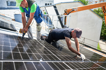 Workers building photovoltaic solar panel system on metal rooftop of house. Men technicians...