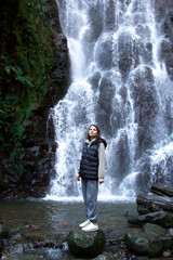 Obraz premium A vertical photo of a woman standing on a rock in front of a cascading waterfall, looking upwards, surrounded by lush greenery, symbolizing reflection and connection with nature