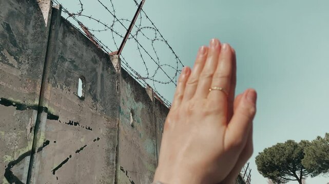 Hopelessness and disappointment of human soul, lost of freedom because of crime commitment, human hands against blue sky and wall with barbed wire background praying for pardon