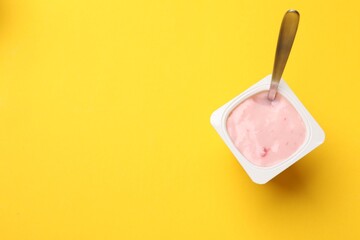 Delicious yogurt in plastic cup with spoon on yellow background, top view. Space for text