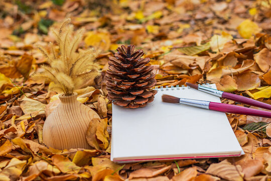 A wooden vase, a pine cone, a blank notebook, and paintbrushes lie on golden autumn leaves