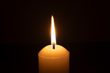 A single burning candle flame or light glowing on a big white candle isolated on black or dark background on table in church for Christmas, funeral or memorial service