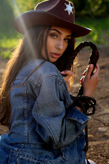 A young woman in a cowboy hat, denim jacket, and boots holding a whip and posing on cracked earth, with a dramatic western-style backdrop.