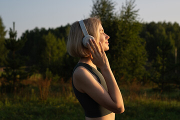 Young woman listening to music and doing breathing exercises in the fresh air. A thin girl meditates while standing against the background of nature. Sporty woman warming up before a morning workout.