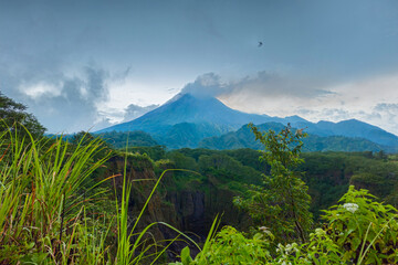 Fototapeta premium Panorama of Mount Merapi from a close distance with rows of clouds around it in the afternoon. Landscape of active volcano in detail from south side with rocks and ravine in front