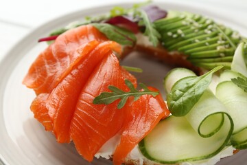 Delicious bagel with salmon, cream cheese, cucumber and avocado on white wooden table, closeup