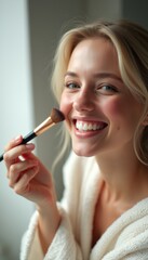 Obraz premium A cheerful young woman with blonde hair smiles brightly while applying blush with a makeup brush, radiating confidence and joy. Her soft, natural beauty is enhanced by the cozy atmosphere, showcasing
