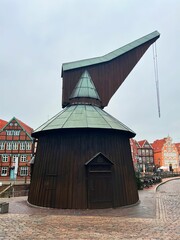 A historic wooden crane with a copper roof stands on a cobblestone square, surrounded by charming half-timbered buildings. The overcast sky adds a nostalgic atmosphere to the scene.