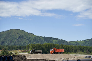 MANZHEROK, ALTAI REPUBLIC, RUSSIA - July 19, 2021: Construction site with trucks on dirt mound under blue sky near verdant hills and lush greenery.