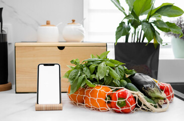 Hand with white screen phone near fresh vegetables and fruits in a reusable bag in the modern kitchen. Food delivery. Grocery shopping online. Eco-Friendly Shopping. The idea of a healthy lifestyle