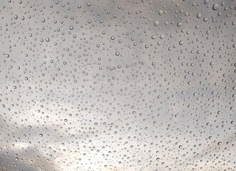 Stunning macro shot of water droplets on glass, showcasing a captivating texture against a blurred cloudy sky. Ideal for backgrounds, websites, or design projects needing a tranquil, atmospheric feel.
