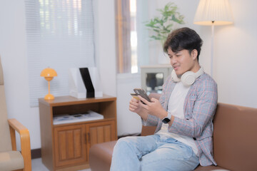 Happy Asian male freelancer sitting on a comfortable sofa at home, holding a credit card and using a smartphone for online shopping, enjoying a seamless e-commerce experience