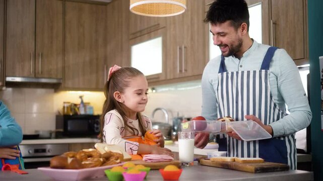 A young father and two small children pack lunch boxes together in the kitchen at home before going to kindergarten.