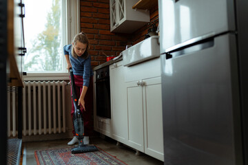 Girl using cordless vacuum cleaner while cleaning kitchen carpet in a modern apartment, focused on...