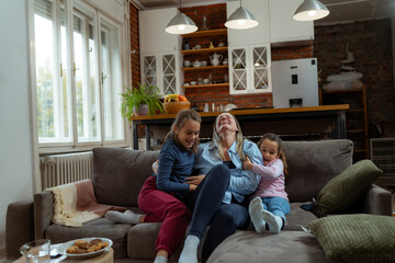 Two daughters are tickling their mother on the sofa in their living room, enjoying a playful and...