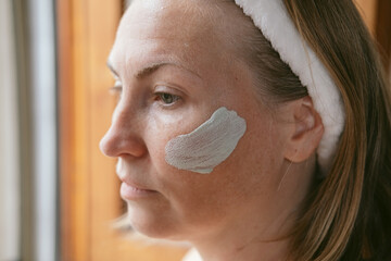 beauty skincare woman applies a cleansing clay mask to her face near the mirror facial skin care...