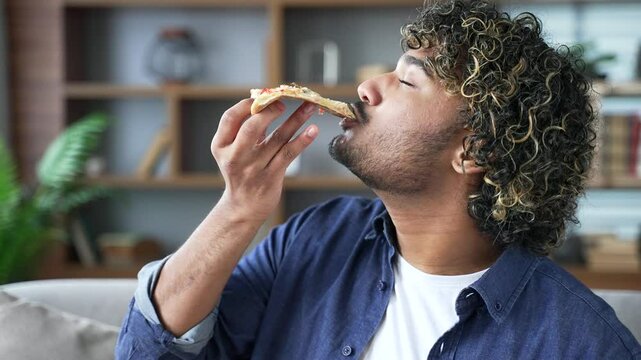 Cheerful satisfied young man enjoys eating pizza sitting on sofa in living room in apartment. Smiling handsome man is happy about fast delivery of food. Ordering food to take home. Side view. Close up