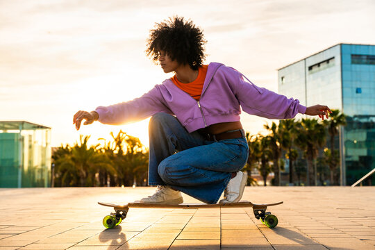 Beautiful young black woman with curly afro hair portrait outdoors - Modern cool female wearing stylish urban clothing strolling in the city with longboard - Powered by Adobe