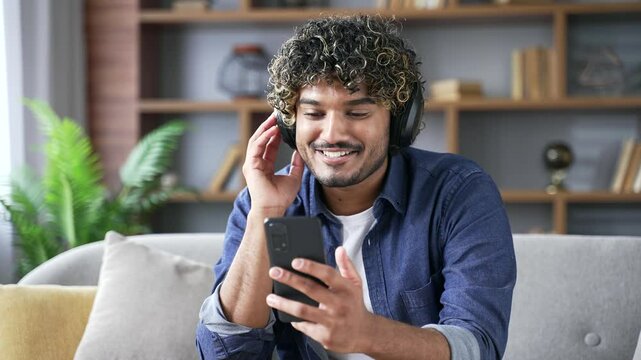 Joyful handsome man wearing wireless headphones listening to music using mobile phone sitting on sofa in living room at home. Happy young adult male turns on a playlist and enjoys favorite song