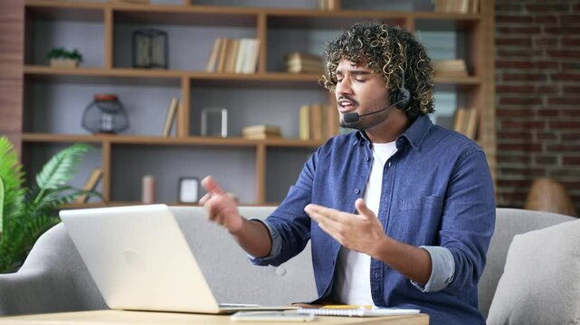 Angry excited young businessman in headset arguing on video call using a laptop computer sitting on sofa in living room at home. Entrepreneur having emotional argument during online business meeting