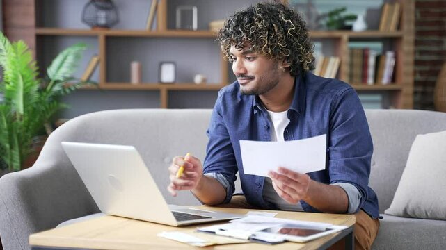 Happy handsome man dealing with household finances using website or app on laptop sitting at home office. Young adult male holds a utility bill and forms payments online by entering data on computer