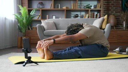 Young handsome man practices gymnastics watching online course on phone sitting on mat on floor in living room at home. Sporty male doing stretching exercises listening to instructor via video call