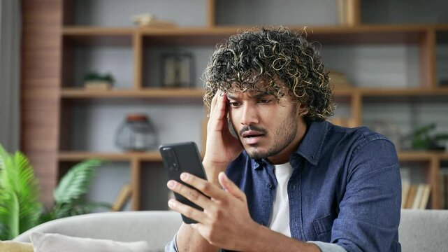 Frustrated young man reads bad news on mobile phone sitting on the sofa in living room at home. Worried depressed adult handsome male is sad while reviewing negative message on smartphone. Close up