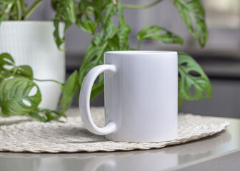 White blank coffee mug on grey table over blured green plant and kitchen interior, mockup