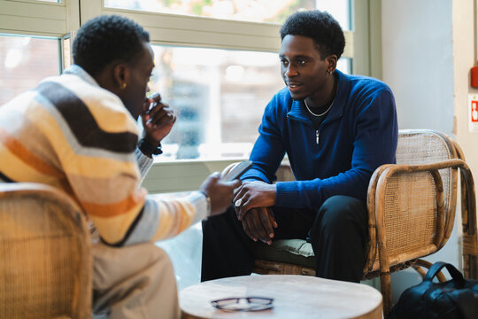 Two black friends seated on wicker chairs, smiling and enjoying a moment of laughter while engaging in an animated conversation in a cozy cafe with a bright, inviting interior.