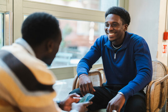 Two black friends seated on wicker chairs, smiling and enjoying a moment of laughter while engaging in an animated conversation in a cozy cafe with a bright, inviting interior.