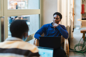 A young black man seated on a wicker chair with a laptop, appearing thoughtful and reflective, in a brightly lit cafe with a modern and inviting atmosphere enhanced by large glass windows.