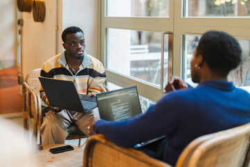 Two young black men actively collaborating on laptops while seated on wicker chairs near large windows in a modern cafe, engaged in an animated exchange of ideas with expressive gestures.