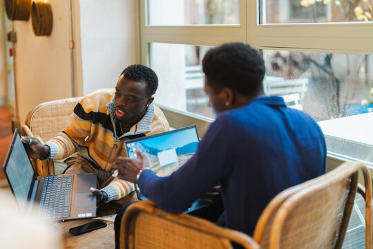 Two young black men actively collaborating on laptops while seated on wicker chairs near large windows in a modern cafe, engaged in an animated exchange of ideas with expressive gestures.
