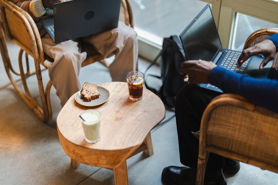 A close-up of two individuals seated in a cafe with laptops on their laps, a wooden table in the center displaying a slice of cake, an iced drink, and a latte