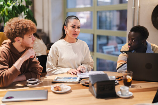 A diverse group of young adults engages in a lively discussion at a cafe table with open laptops, notebooks, and cups of coffee, surrounded by a cozy and modern setting with natural light. - Powered by Adobe