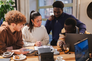 A group of young adults collaborates at a cafe table while reviewing a smartphone, notebooks, and laptops, engaging in creative teamwork within a modern and welcoming environment.