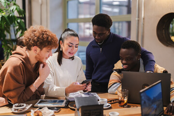 A group of young adults collaborates at a cafe table while reviewing a smartphone, notebooks, and laptops, engaging in creative teamwork within a modern and welcoming environment.