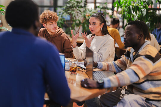 A group of young adults engages in a lively discussion at a cafe table, surrounded by laptops, notebooks, and coffee, with natural light and greenery complementing the collaborative atmosphere.