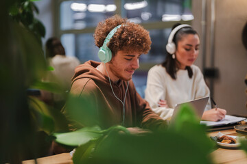 Young individuals with headphones focus on their work at a cafe table, engaging with laptops and notebooks amidst a modern and creative environment with greenery and natural light.