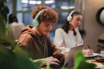 Young individuals with headphones focus on their work at a cafe table, engaging with laptops and notebooks amidst a modern and creative environment with greenery and natural light.