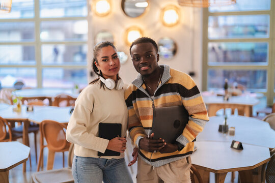 A young man and woman pose confidently with a laptop and tablet in a stylish, modern cafe setting, showcasing a collaborative and creative atmosphere enhanced by warm lighting.