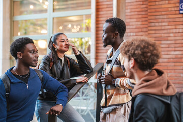 A diverse group of four young adults gathers near a modern building, engaged in a lively conversation while carrying backpacks, showcasing collaboration and community in a stylish setting.