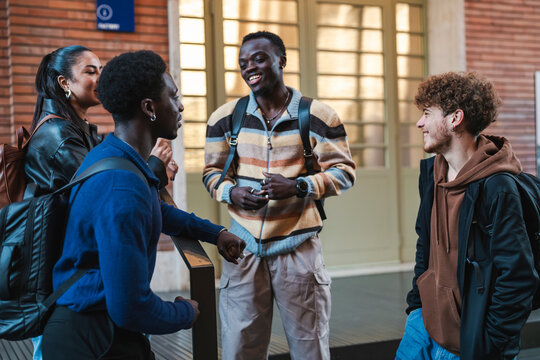 Four young adults engage in a friendly conversation while standing outside a modern building, wearing casual outfits and backpacks, emphasizing teamwork and connection in a relaxed urban setting. - Powered by Adobe