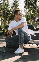 Trendy young man squatting by parked motorbike while using drone controller in nature