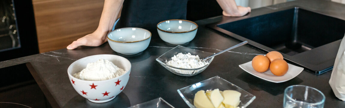 Banner of unrecognizable young pastry chef is arranging bowls of flour, sugar, butter, and eggs on a sleek, dark countertop in a contemporary kitchen, ready to begin baking