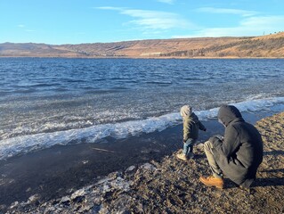Obraz premium A father and his two-year-old son share a joyful moment playing at the frozen shoreline of a mountain lake on a winter day. Snow surrounds them, creating a serene and picturesque scene of family conne