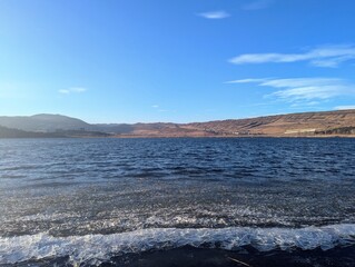 A stunning view of a mountain lake on a bright, sunny day, with ice still clinging to the shoreline. The sparkling ice reflects the sunlight, creating a mesmerizing contrast with the surrounding lands