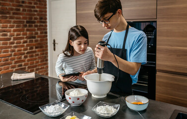Teenage brother mixing ingredients for a cake using hand blender while curious sister looking...
